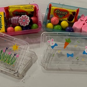 May include: Two clear plastic containers with lids. One container has a pink lid and contains crayons, candy, and a small flower. The other container has a blue lid and contains pink marshmallow Peeps and candy. Both containers have a decorative design on the bottom.