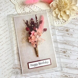 May include: A birthday card featuring a bouquet of dried flowers in shades of pink, burgundy, and cream. The card is encased in a clear plastic box, with the words "Happy Birthday" displayed on a rectangular label.