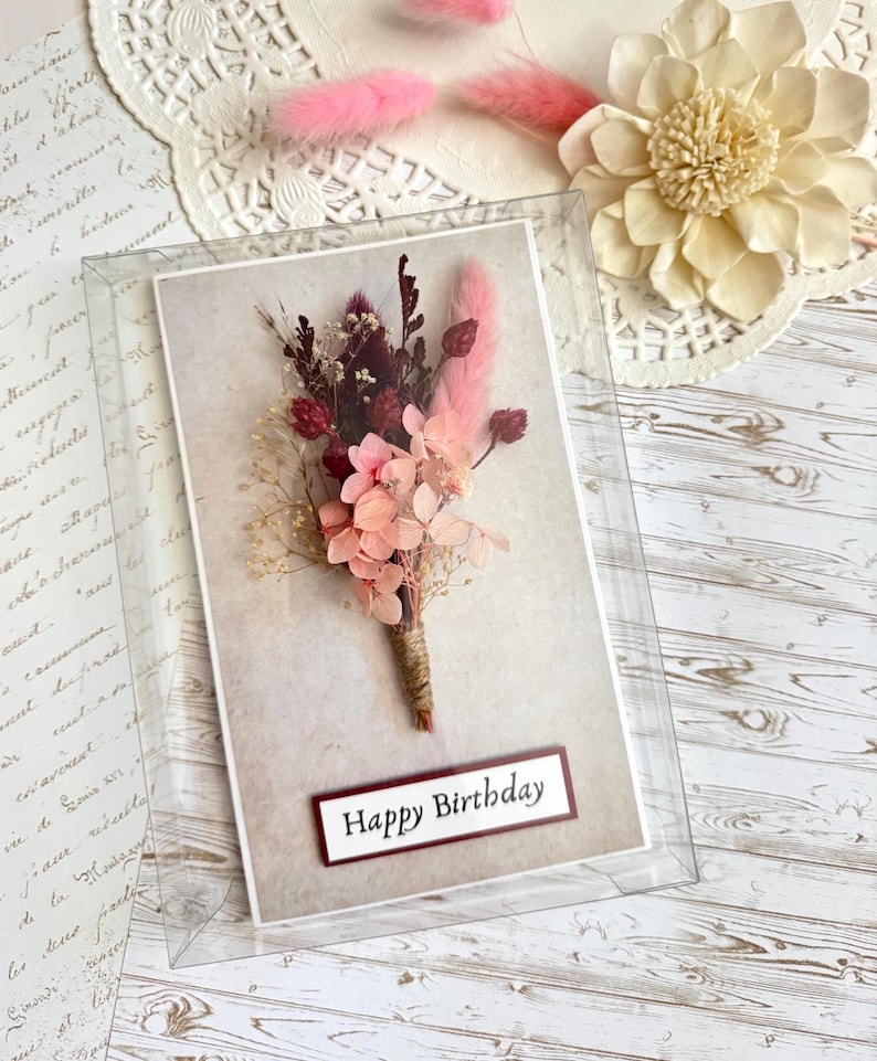May include: A birthday card featuring a bouquet of dried flowers in shades of pink, burgundy, and cream. The card is encased in a clear plastic box, with the words "Happy Birthday" displayed on a rectangular label.