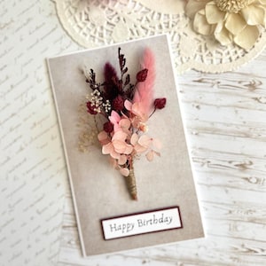 May include: A greeting card with a floral arrangement. The card has a beige background with a small bouquet of pink and burgundy dried flowers tied with twine. The words "Happy Birthday" are printed on a rectangular label.