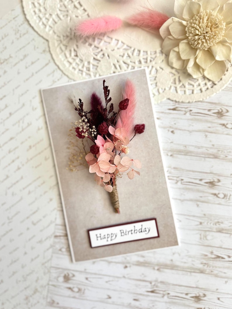 May include: A greeting card with a floral arrangement. The card has a beige background with a small bouquet of pink and burgundy dried flowers tied with twine. The words "Happy Birthday" are printed on a rectangular label.