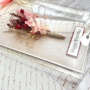 May include: A birthday card featuring a bouquet of dried flowers in shades of pink and burgundy, wrapped with twine. The card is in a clear plastic sleeve with a "Happy Birthday" label.
