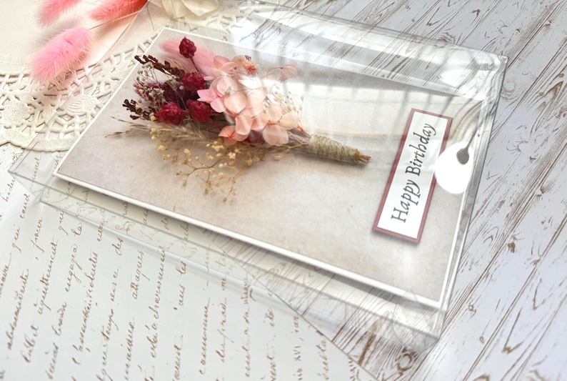 May include: A birthday card featuring a bouquet of dried flowers in shades of pink and burgundy, wrapped with twine. The card is in a clear plastic sleeve with a "Happy Birthday" label.