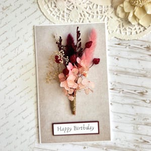May include: A birthday card featuring a bouquet of dried flowers in shades of pink, burgundy, and cream. The card has the words "Happy Birthday" in a rectangular frame. The background is a neutral color.