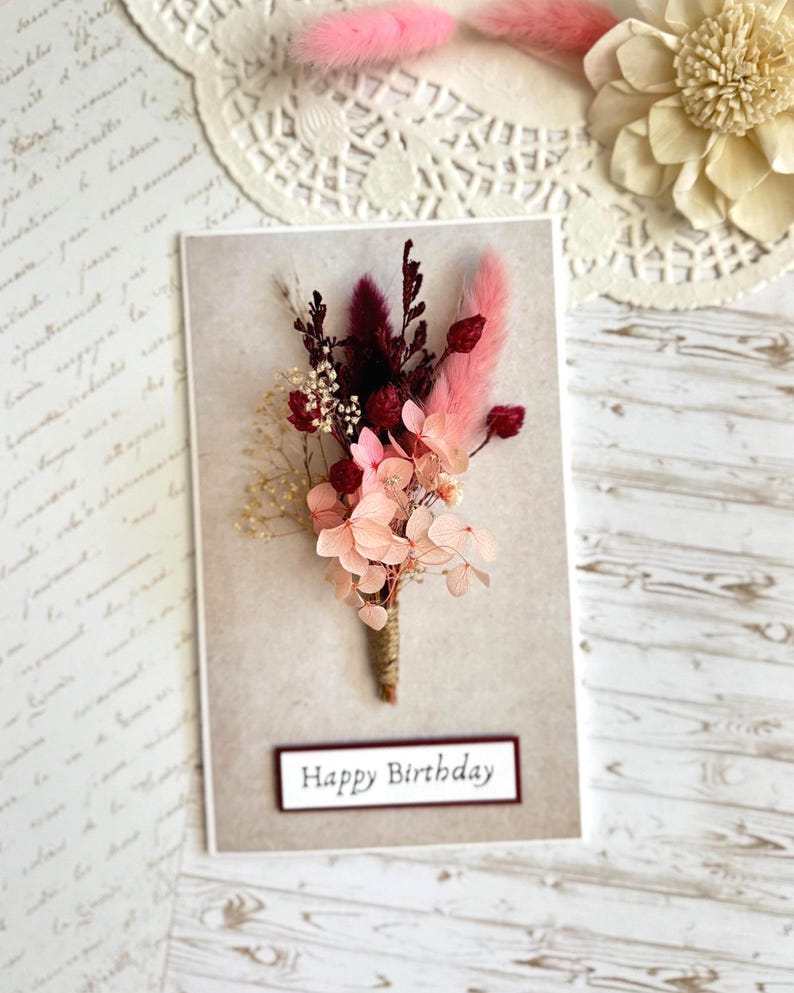May include: A birthday card featuring a bouquet of dried flowers in shades of pink, burgundy, and cream. The card has the words "Happy Birthday" in a rectangular frame. The background is a neutral color.