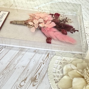 May include: A floral arrangement in a clear acrylic box. The bouquet features pink and burgundy dried flowers, with a pink feather accent. A rectangular label with the words "Happy Birthday" is visible. The arrangement is on a light wood-grain surface.