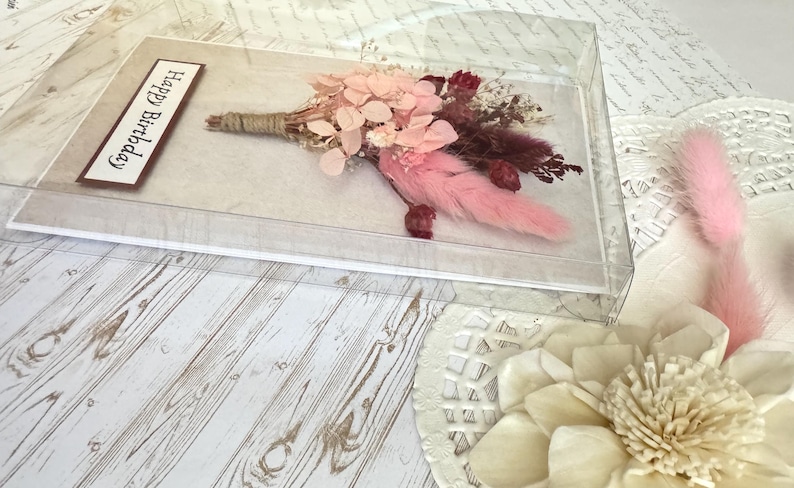 May include: A floral arrangement in a clear acrylic box. The bouquet features pink and burgundy dried flowers, with a pink feather accent. A rectangular label with the words "Happy Birthday" is visible. The arrangement is on a light wood-grain surface.