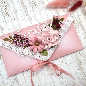 May include: A pink envelope decorated with paper flowers, lace, and a ribbon. The envelope is adorned with a cluster of pink and burgundy paper flowers, white lace trim, and a delicate pink ribbon tied in a bow.