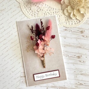 May include: A greeting card with a floral arrangement and the text "Happy Birthday." The card features a bouquet of dried flowers in shades of pink, burgundy, and cream, tied with twine. The background is a neutral beige.