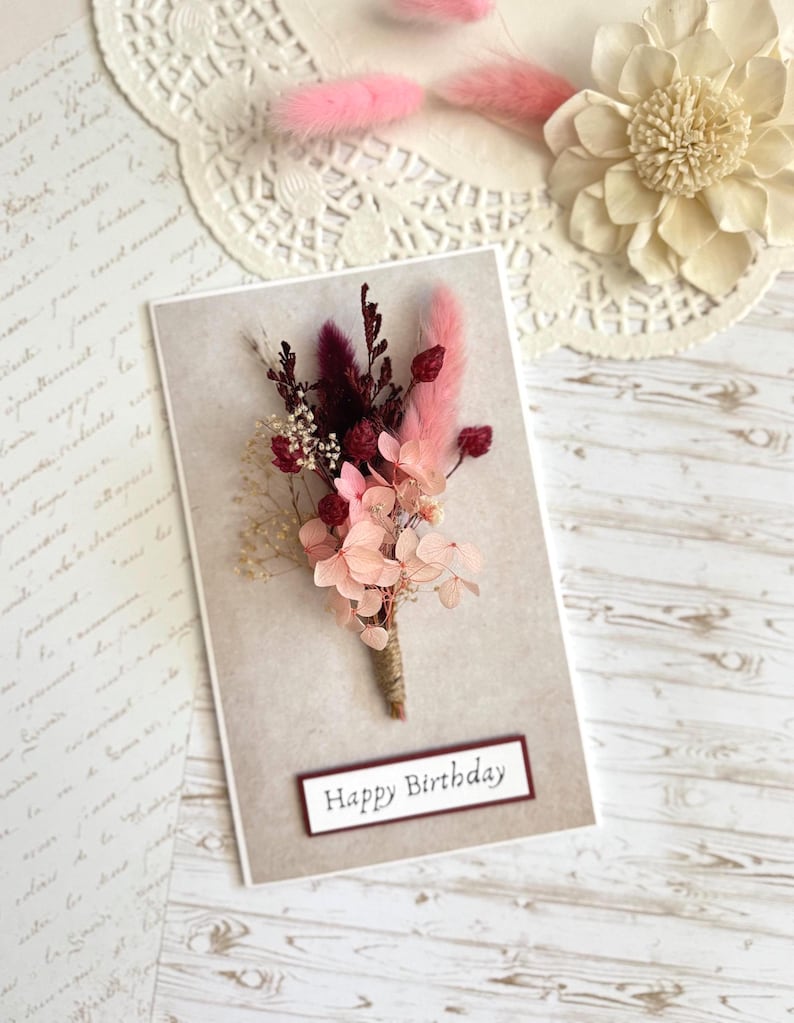 May include: A greeting card with a floral arrangement and the text "Happy Birthday." The card features a bouquet of dried flowers in shades of pink, burgundy, and cream, tied with twine. The background is a neutral beige.