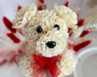 Könnte beinhalten: Dekorative Hundefigur aus cremefarbenen Nelken, mit schwarzen Perlenaugen und -nase. Ein rotes Schleifenband schmückt die Basis, mit roten und weißen floralen Akzenten. Der Hund steht auf einer weißen Spitzendeckchen.