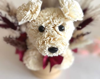 Könnte beinhalten: Eine cremefarbene Blumenskulptur eines Hundes mit schwarzen Perlenaugen und einer schwarzen Nase. Der Hund hat Schlappohren und ist mit einer bordeauxfarbenen Schleife verziert. Trockenblumen in Creme-, Bordeaux- und Rosatönen umgeben den Hund.