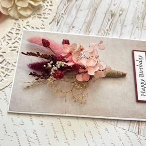 May include: A greeting card with a floral arrangement. The card features a bouquet of dried flowers in shades of pink, burgundy, and cream, tied with twine. The words "Happy Birthday" are printed on a rectangular label.