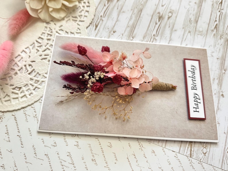 May include: A greeting card with a floral arrangement. The card features a bouquet of dried flowers in shades of pink, burgundy, and cream, tied with twine. The words "Happy Birthday" are printed on a rectangular label.