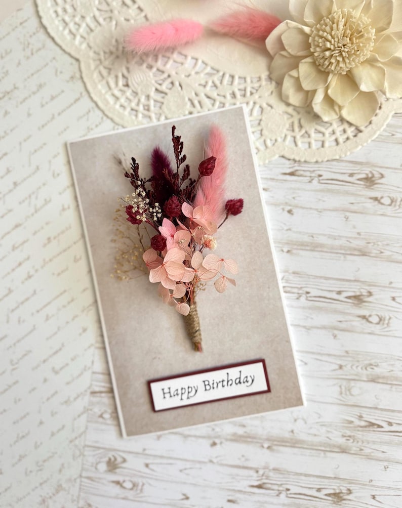 May include: A greeting card with a floral arrangement and the text "Happy Birthday." The arrangement features pink and burgundy dried flowers and is tied with twine. The card has a neutral background.