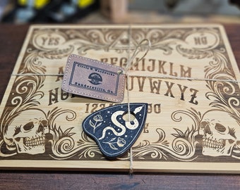 Könnte beinhalten: Ein Ouija-Brett aus Holz mit einer schwarzen und weißen Planchette mit einem Schlangenmotiv. Das Brett hat ein traditionelles Layout mit den Buchstaben A-Z, den Zahlen 0-9 und den Wörtern "Ja", "Nein" und "Auf Wiedersehen".