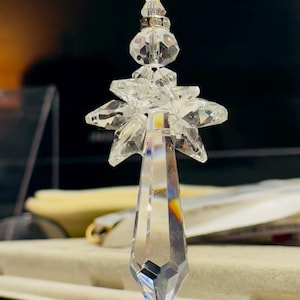 May include: A clear crystal prism ornament with a starburst design. The ornament has a faceted, elongated teardrop shape, with a series of clear crystal beads above the starburst. The ornament is suspended from a clear string.