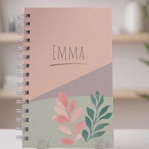 Puede incluir: Un cuaderno de espiral de 120 páginas con un fondo geométrico en rosa empolvado y verde salvia. La portada muestra el nombre EMMA en una fuente sans-serif sencilla sobre una línea horizontal, con ilustraciones de hojas rosas y verde oscuro.