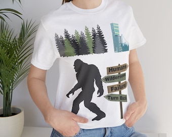 May include: White t-shirt featuring a black silhouette of Bigfoot walking, with a forest and city skyline graphic. Directional signs read "Humans," "Wilderness," "Failure," and "Turn Back." The shirt is a casual, short-sleeved design.