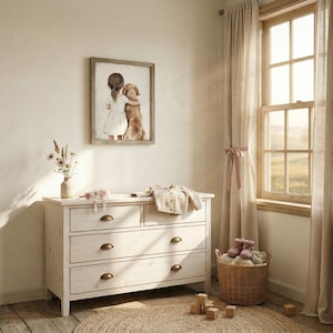 May include: A light-colored wooden dresser with four drawers and brass-colored handles. A framed picture of a girl hugging a dog hangs on the wall above the dresser. A small vase of flowers and a baby sweater sit on top of the dresser. A woven basket and a rug are also in the room.