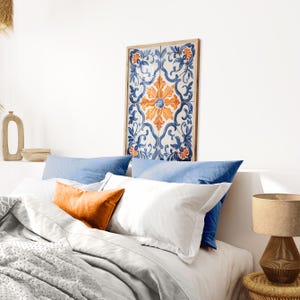 May include: A white bed with blue and orange pillows and a white blanket. A framed artwork with a blue and orange floral pattern hangs above the bed.