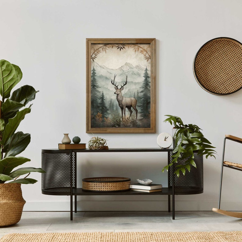 May include: A framed print of a watercolor painting depicting a deer standing in a forest with mountains in the background. The print is in a wooden frame with a light brown finish.