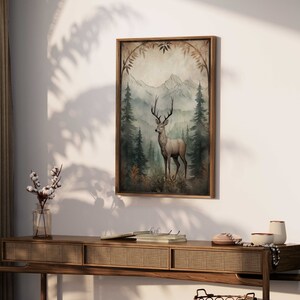 May include: A framed print of a watercolor painting depicting a deer standing in a forest with mountains in the background. The deer is brown with a white underbelly and has large antlers. The forest is green and the mountains are blue and purple.