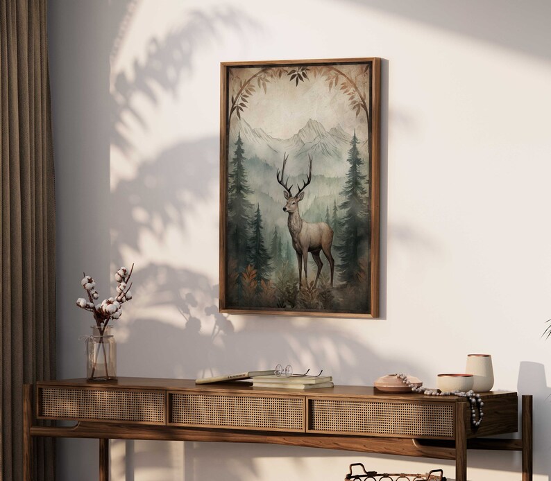 May include: A framed print of a watercolor painting depicting a deer standing in a forest with mountains in the background. The deer is brown with a white underbelly and has large antlers. The forest is green and the mountains are blue and purple.