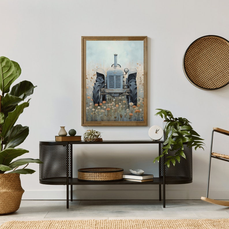 May include: A framed painting of a blue vintage tractor in a field of wildflowers. The tractor is facing the viewer and is surrounded by a variety of flowers in shades of orange, pink, and yellow. The painting is set against a white wall and is displayed on a black metal shelf with a woven basket and books.