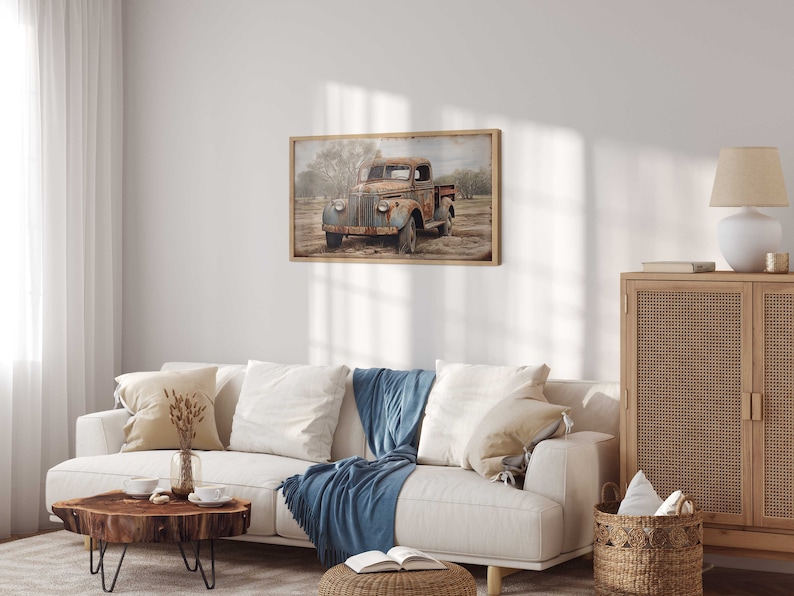 May include: A vintage, rusty blue pickup truck is parked in a field with trees in the background. The truck is framed in a wooden frame and hangs on a white wall in a living room setting.