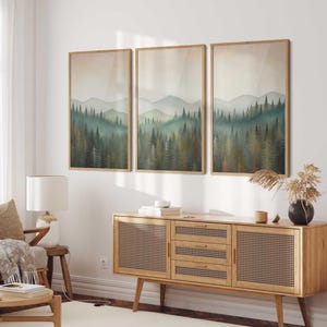 May include: Three framed landscape paintings of a forest with a misty background. The paintings are hung on a white wall above a wooden cabinet with a woven rattan front.