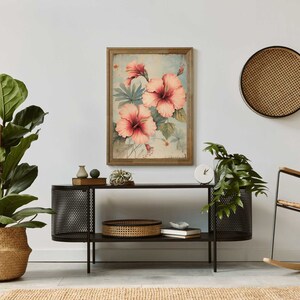 May include: A framed print of pink hibiscus flowers with green leaves on a light blue background. The print is hanging on a white wall above a black metal console table with a woven basket and books on the shelf.