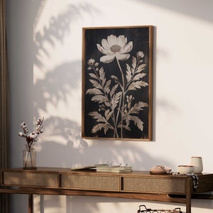 May include: A black and white botanical print of a single flower with leaves on a dark wood background. The print is framed in a dark wood frame.