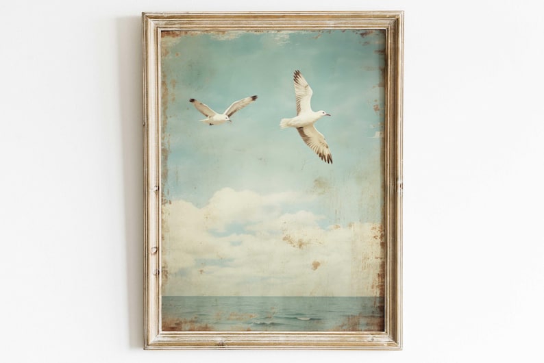 Puede incluir: Una impresi&oacute;n enmarcada de estilo vintage con dos gaviotas blancas volando contra un cielo azul con nubes blancas. El oc&eacute;ano es visible en la parte inferior de la imagen.