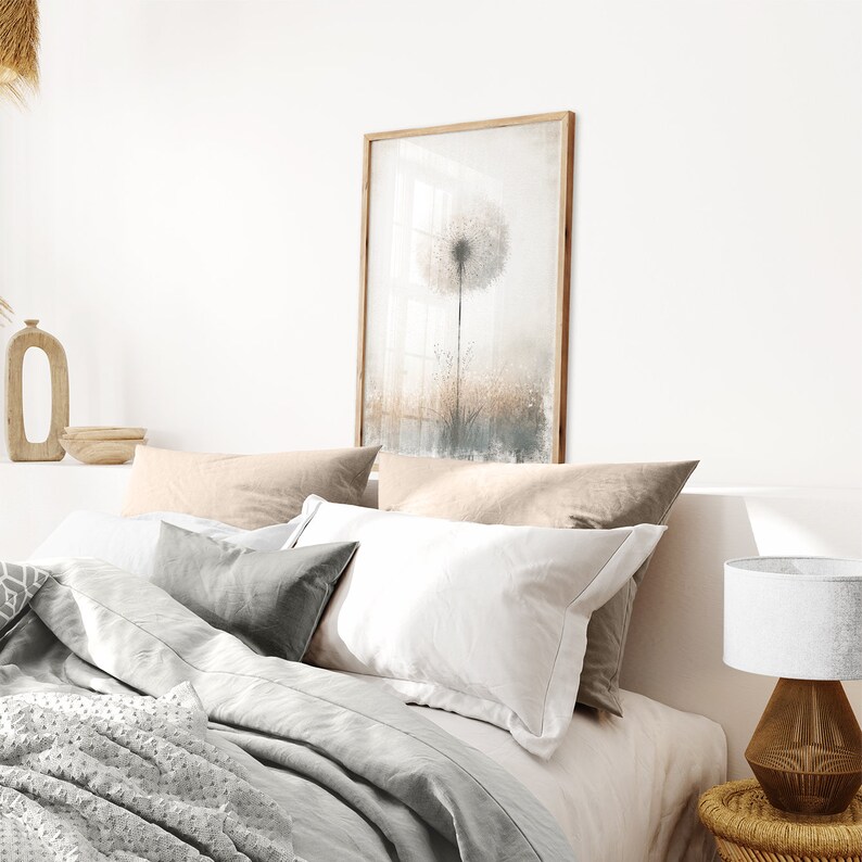 May include: A bedroom with a white bed, a grey blanket, and a framed print of a dandelion. The bed has a white pillow, a grey pillow, and two beige pillows. There is a lamp on a small table next to the bed.