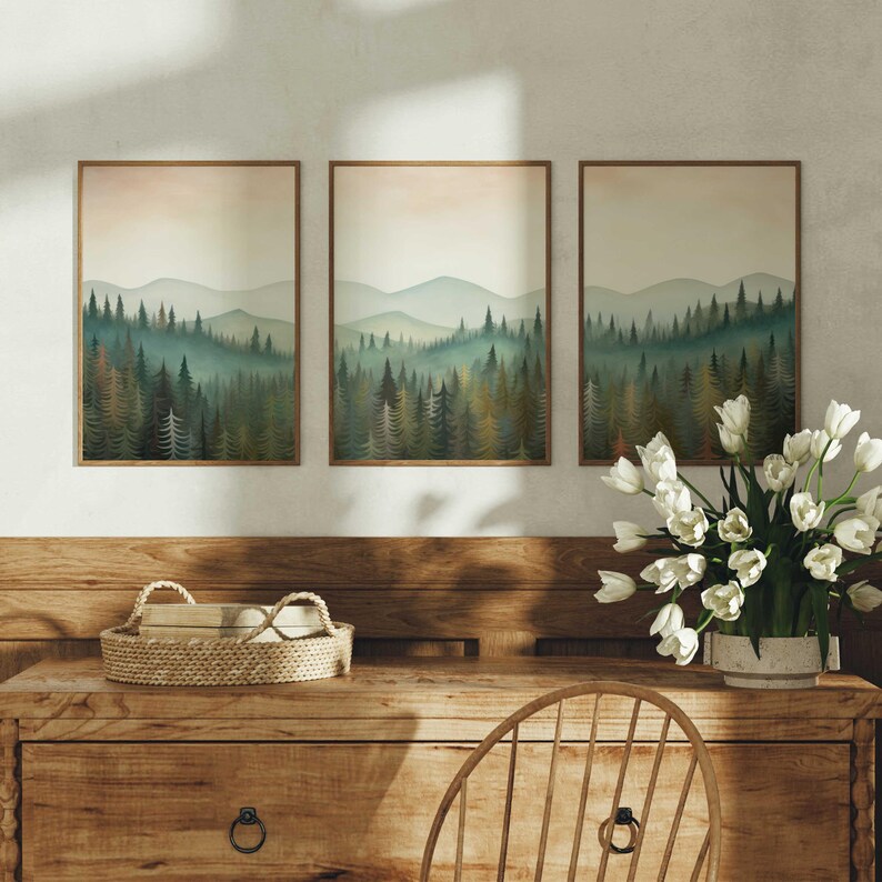 May include: Three framed prints of a misty forest scene with a row of trees in the foreground and a mountain range in the background. The prints are hung on a white wall above a wooden dresser.
