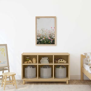 May include: A light wood bookshelf with six cubbies, each containing a storage basket or decorative items. The bookshelf is in a room with a white wall, a wooden easel with a drawing of a sailboat and sun, a small wooden stool, and a wooden bed with a checkered patterned comforter.