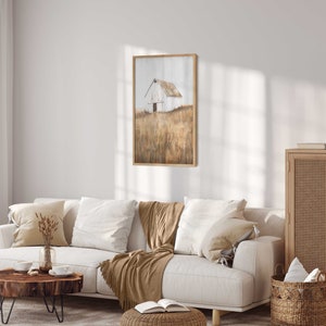 May include: A framed painting of a white barn in a field of tall grass. The barn has a brown roof and a single door. The grass is a muted brown and yellow color. The painting is in a light wood frame.