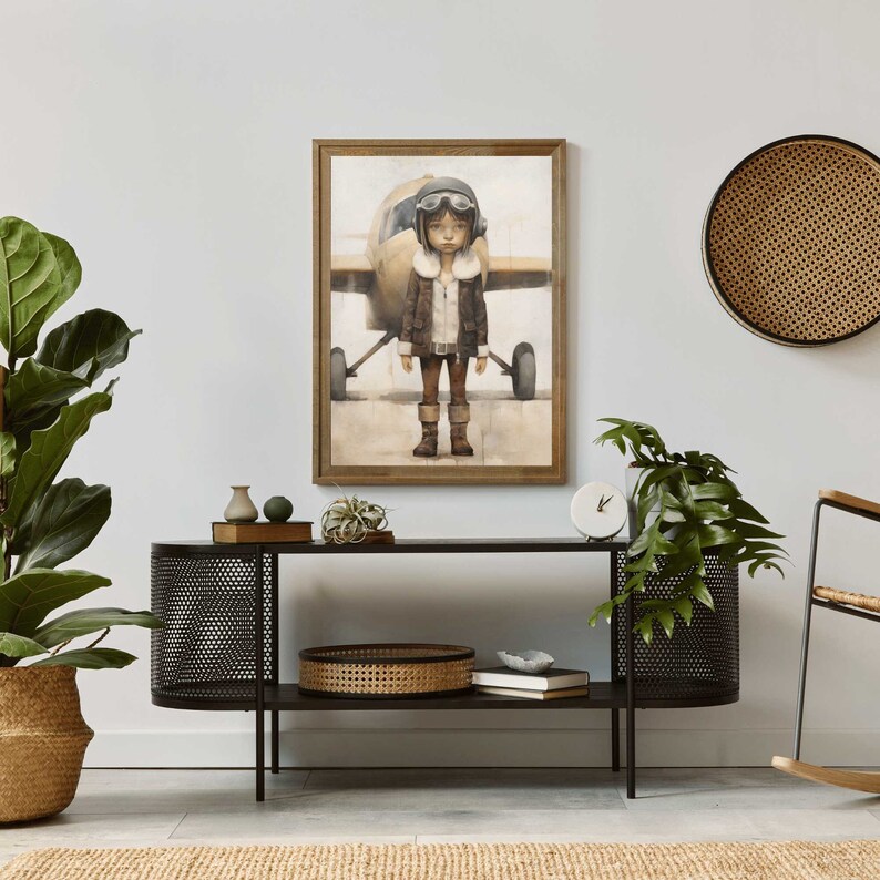 Op de afbeelding: Een ingelijst schilderij van een jong persoon die een bruin leren jack en een vliegeniersbril draagt en voor een klein vliegtuig staat. Het schilderij is geplaatst tegen een witte muur met een zwart metalen plank en een rieten mand op de plank.