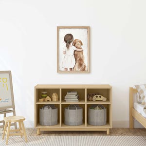 May include: A framed watercolor print depicts a girl in a white dress hugging a golden retriever. The artwork hangs above a wooden storage unit with gray fabric bins. A child's bed with a checkered pillow and a small easel with a drawing are also visible.