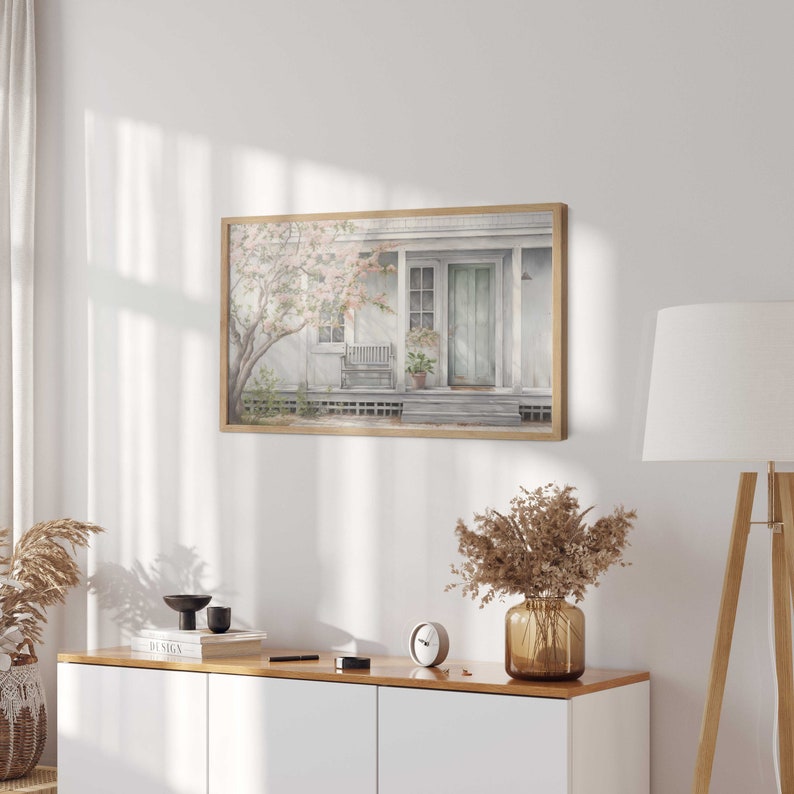 K&ouml;nnte beinhalten: Ein Aquarellbild eines wei&szlig;en Hauses mit einer hellblauen T&uuml;r und einer Veranda mit einer Bank. Das Haus ist von B&auml;umen mit rosa Bl&uuml;ten umgeben.