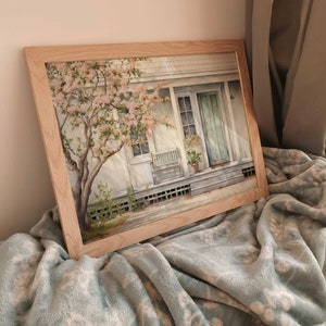 K&ouml;nnte beinhalten: Ein gerahmtes Aquarellbild eines wei&szlig;en Hauses mit einer gr&uuml;nen T&uuml;r und einem Baum mit rosa Bl&uuml;ten davor. Das Haus hat eine Veranda mit einem wei&szlig;en Schaukelstuhl und einer Topfpflanze. Das Bild h&auml;ngt auf einer hellblauen Decke.