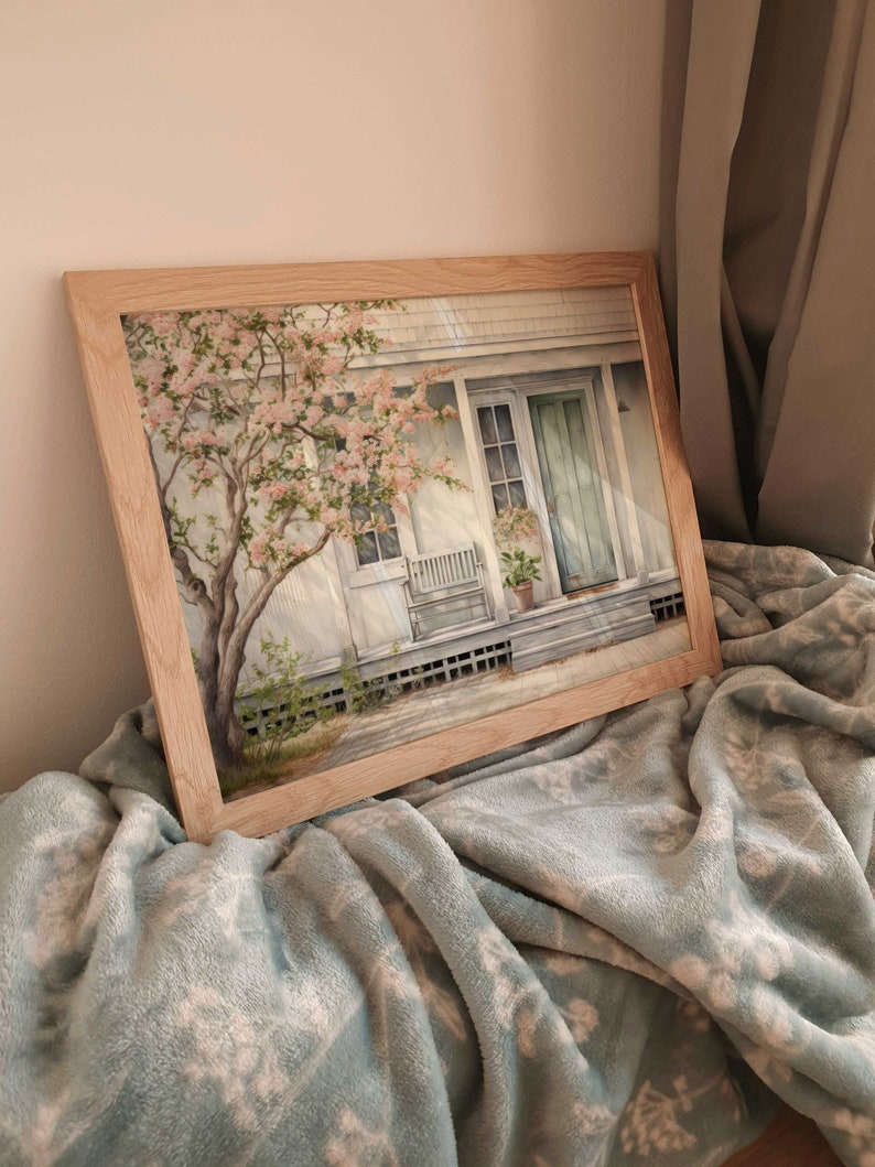K&ouml;nnte beinhalten: Ein gerahmtes Aquarellbild eines wei&szlig;en Hauses mit einer gr&uuml;nen T&uuml;r und einem Baum mit rosa Bl&uuml;ten davor. Das Haus hat eine Veranda mit einem wei&szlig;en Schaukelstuhl und einer Topfpflanze. Das Bild h&auml;ngt auf einer hellblauen Decke.
