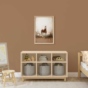 May include: A framed print of a brown horse in a field is displayed above a wooden storage unit with grey fabric bins. A child's easel with a drawing of a sailboat and a small wooden stool are in the foreground. A bed with a checked duvet is on the right.