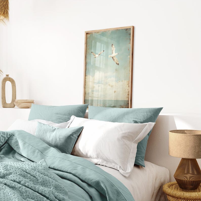 Puede incluir: Un dormitorio con una colcha azul claro y almohadas blancas. Un cuadro enmarcado de dos gaviotas volando sobre un cielo azul y blanco cuelga de la pared sobre la cama.