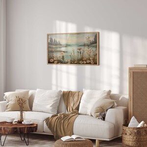 Puede incluir: Un sof&aacute; blanco con cojines beige y una manta de lana marr&oacute;n en una sala de estar con un armario de madera, una l&aacute;mpara y un cuadro enmarcado de un lago con flores.