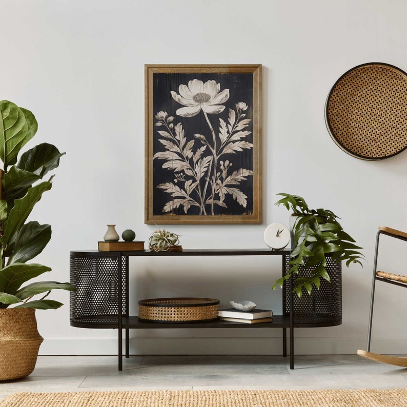 May include: A black metal console table with two shelves. The top shelf has a white vase, a small green plant, and a book. The bottom shelf has a woven basket, a book, and a small white clock. A black and white botanical print hangs on the wall above the table.