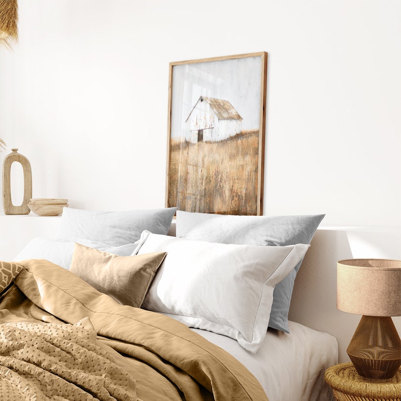 May include: A framed painting of a white barn in a field of tall grass. The barn is painted in a rustic style with a weathered look. The grass is a warm brown color and the sky is a light blue. The painting is framed in a simple wooden frame.