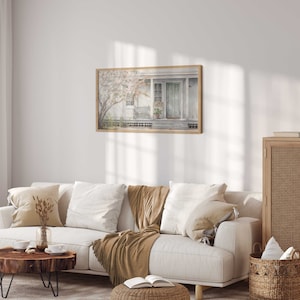 K&ouml;nnte beinhalten: Ein wei&szlig;es Sofa mit braunen &Uuml;berw&uuml;rfen und Kissen in einem Wohnzimmer mit einem gerahmten Aquarellbild eines Hauses mit einer Veranda und einer Lampe auf einem Holzschrank.