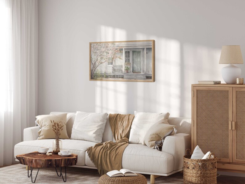 K&ouml;nnte beinhalten: Ein wei&szlig;es Sofa mit braunen &Uuml;berw&uuml;rfen und Kissen in einem Wohnzimmer mit einem gerahmten Aquarellbild eines Hauses mit einer Veranda und einer Lampe auf einem Holzschrank.